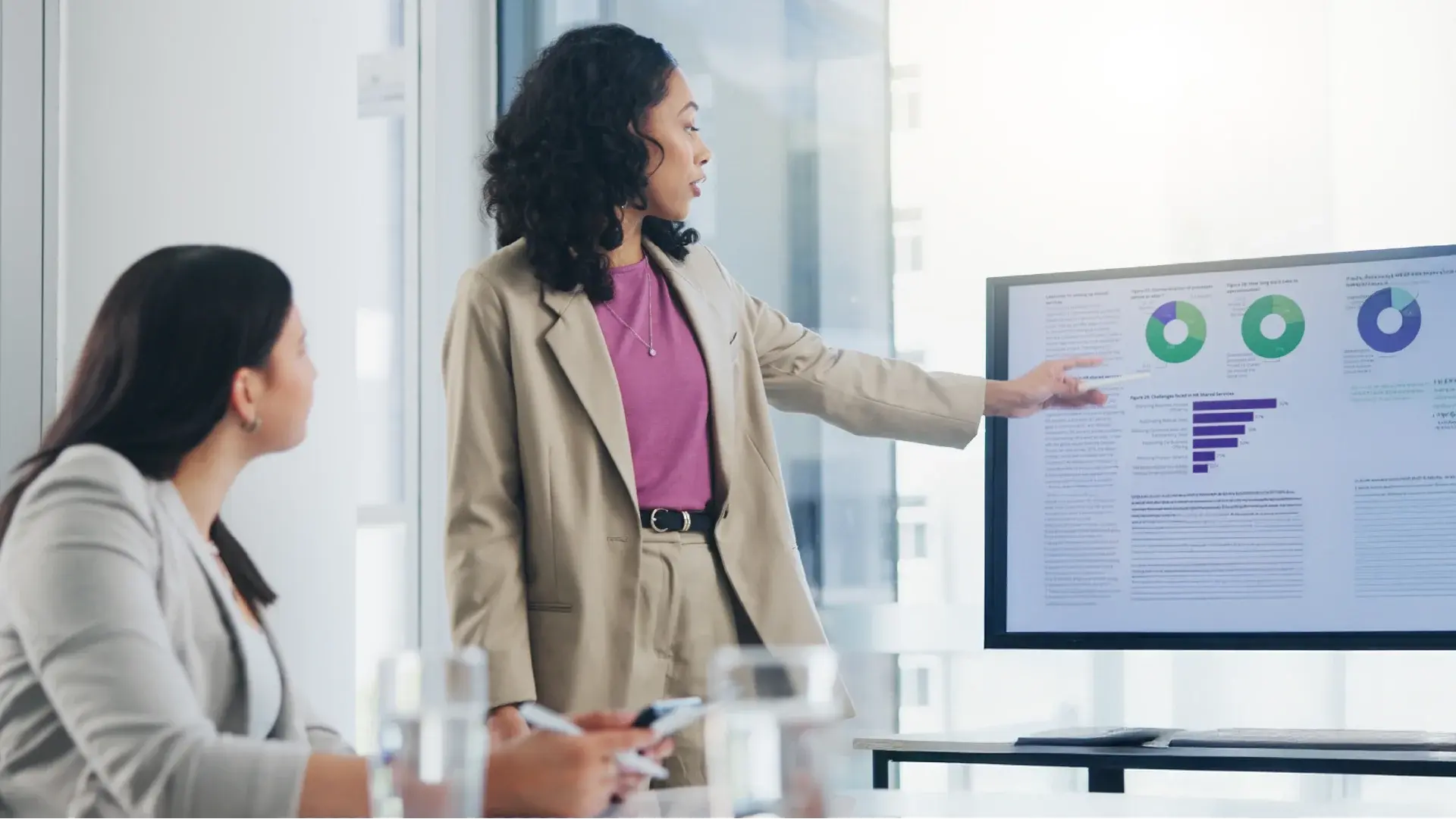 Person standing and pointing at a large monitor displaying charts and graphs during a business presentation, while another person sits at a conference table with documents and a smartphone.